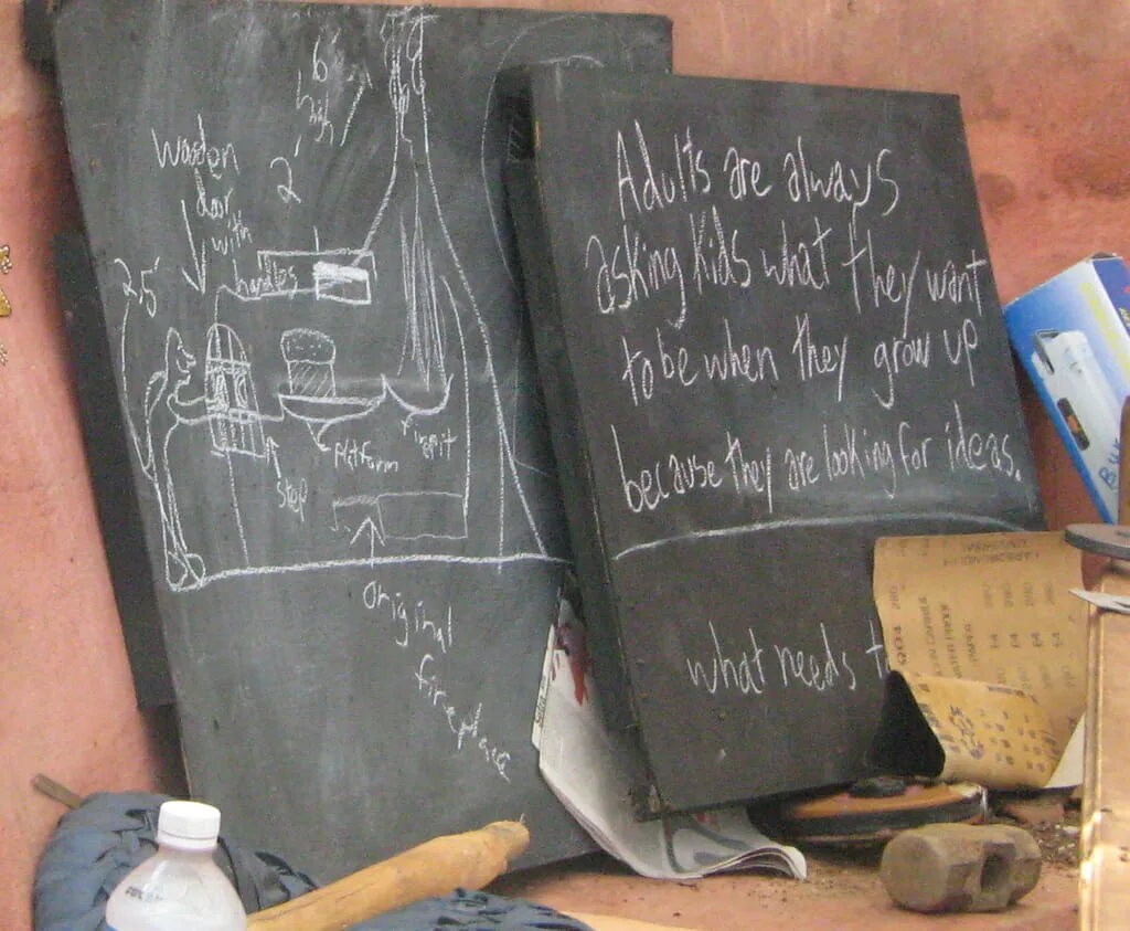 two blackboards sitting outside (it appears) leaning against the side of a stucco house. One has drawings of a fireplace, perhaps planning on how to make it. The other has words, 
